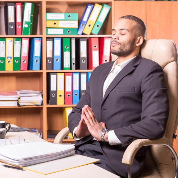 Black man meditating at his work desk, taking time out during a busy day to reduce his stress levels and increase his focus and productivity in the work place